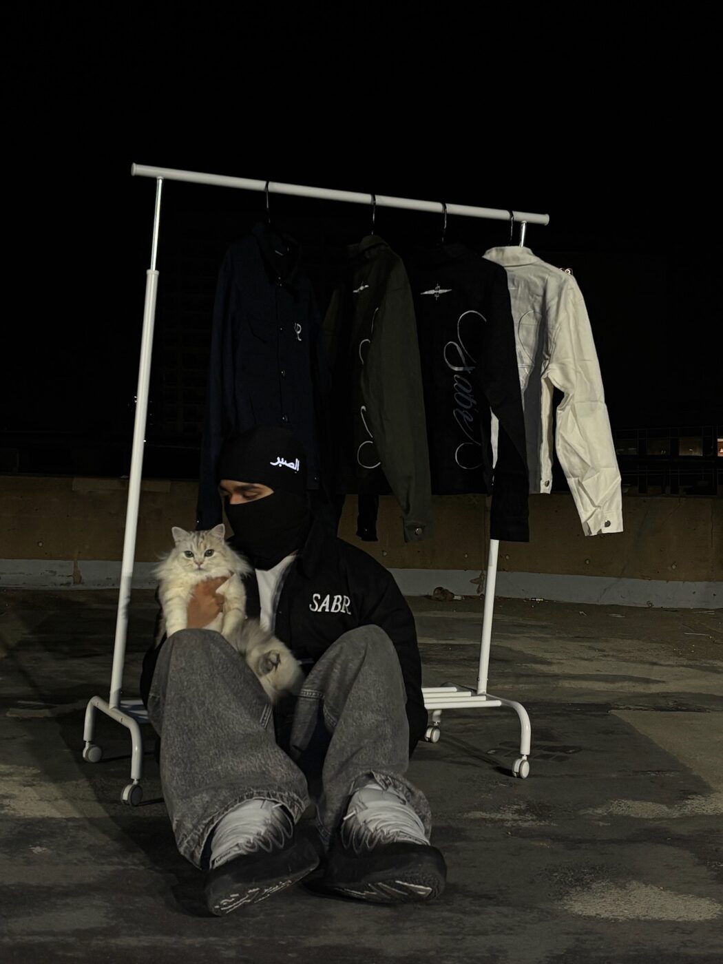 A rail of Sabr jackets on a rooftop at night, a masked figure seated below holding a white cat